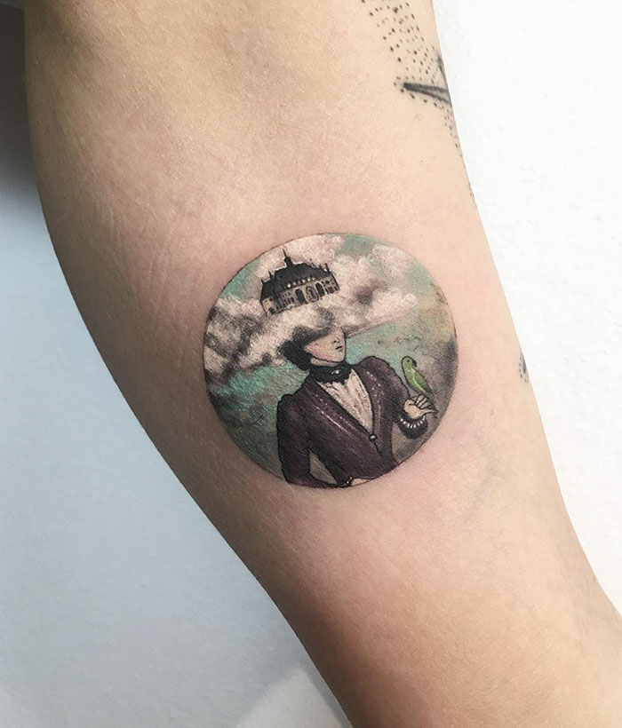 Miniature circle tattoo by Turkish artist Eva Krbdk, depicting a man with clouds and a building on his head, on a forearm.
