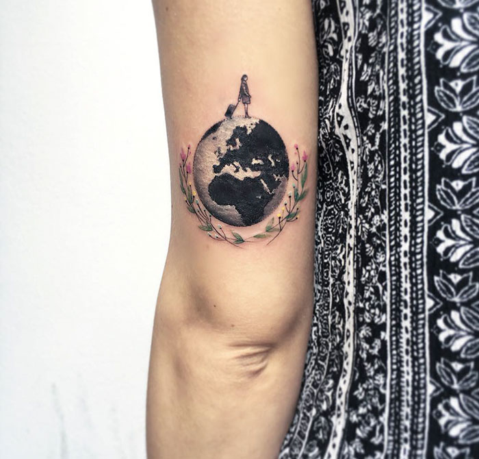 Miniature circle tattoo of Earth with a figure and floral wreath, created by Turkish artist Eva Krbdk on an arm.