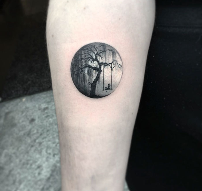 Miniature circle tattoo by Turkish artist, depicting a tree and swing, on a person's arm.