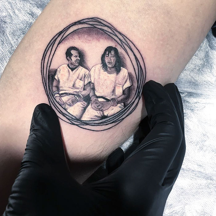 Gloved hand displaying a miniature circle tattoo by Turkish artist Eva Krbdk, featuring two seated figures.