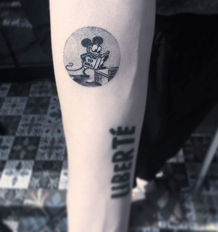 Miniature circle tattoo of a cartoon character reading on an arm with "LIBERTÉ" text below it.