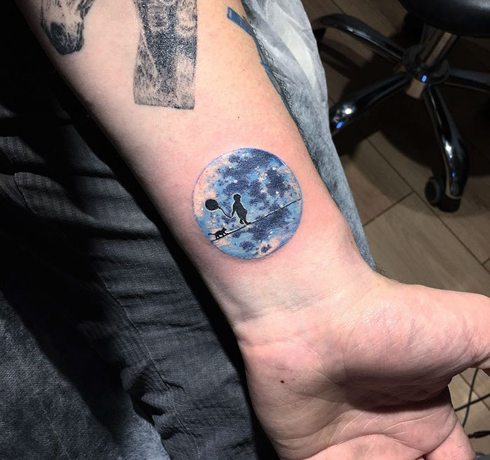 Miniature circle tattoo by Turkish artist Eva Krbdk, featuring a silhouette of a person and a cat inside a cosmic scene.