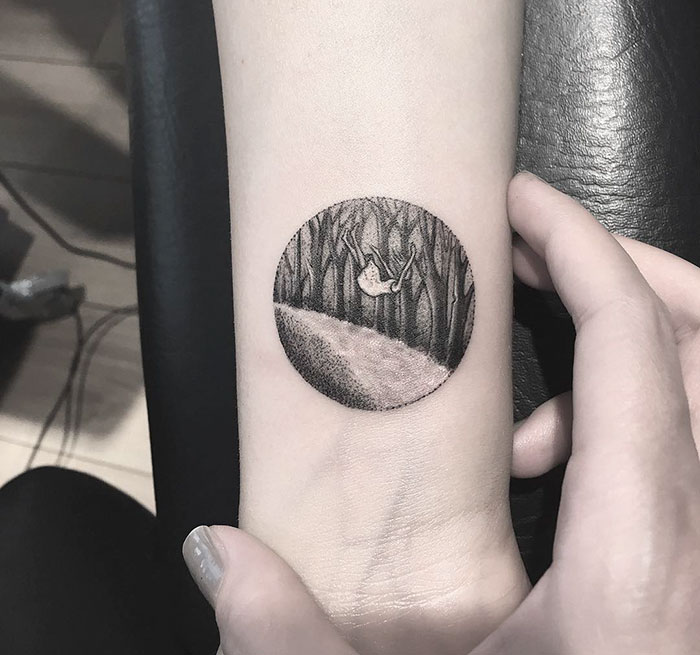 Miniature circle tattoo by Turkish artist Eva Krbdk, featuring a swinging figure inside a forest scene on a person's arm.
