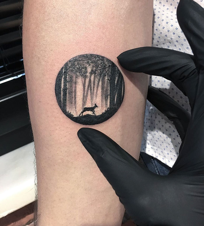 Black-gloved hand holding a miniature circle tattoo depicting a deer in the woods.