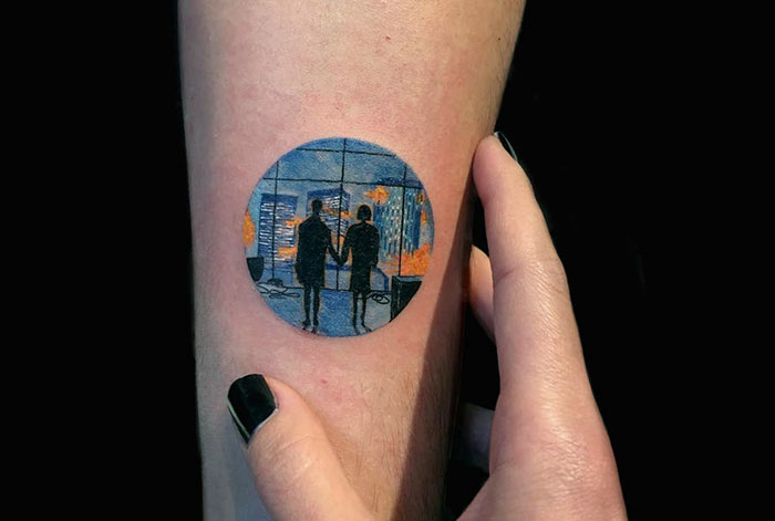 Miniature circle tattoo by Turkish artist Eva Krbdk, depicting silhouettes in a cityscape on forearm.