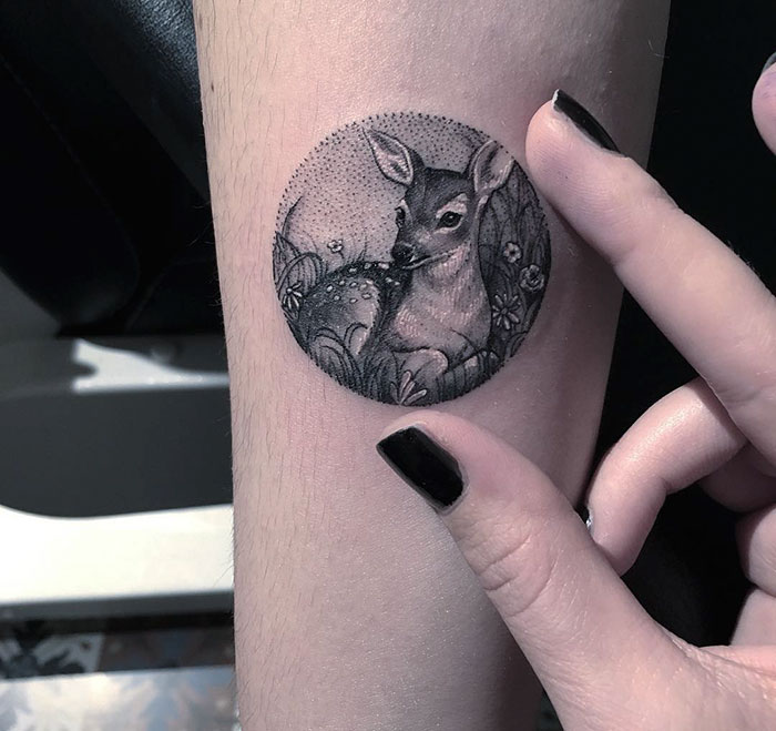 Miniature circle tattoo of a deer on an arm, with a finger pointing to it, by Turkish artist Eva Krbdk.