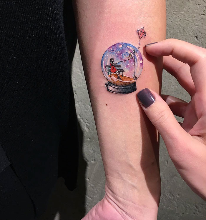 Miniature circle tattoo by Turkish artist Eva Krbdk on forearm, depicting a small scene inside a sphere.