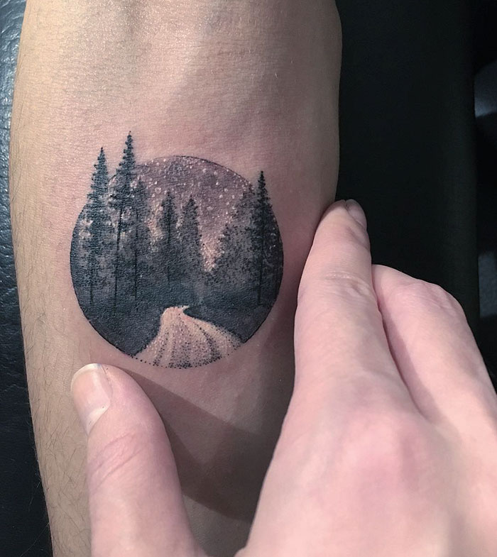Miniature circle tattoo of a serene forest landscape on skin by Turkish artist Eva Krbdk.