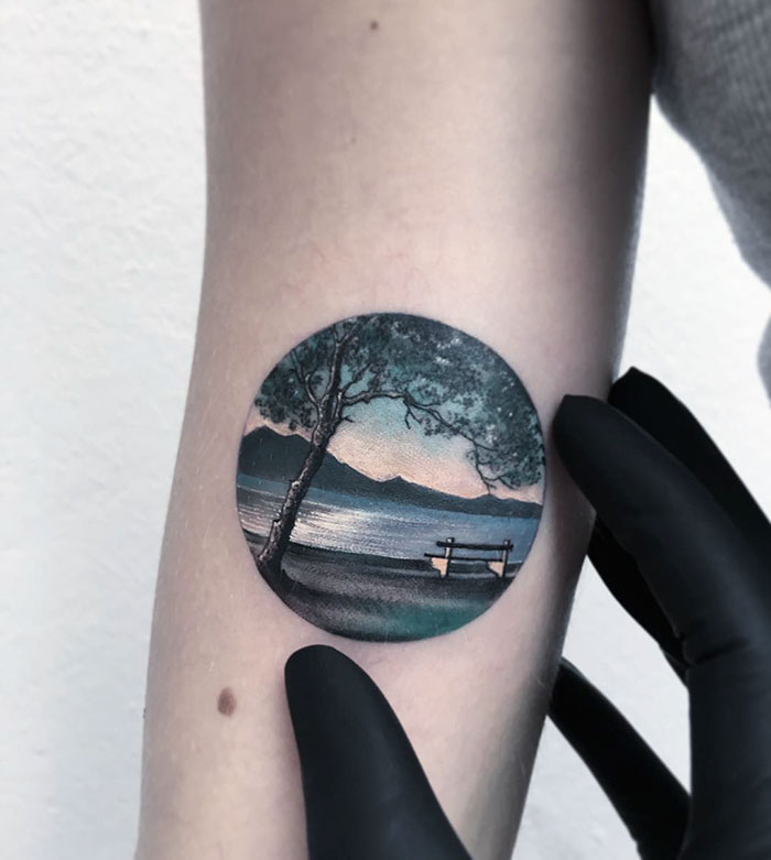 Gloved hand showing a miniature circle tattoo of a serene landscape by Turkish artist.