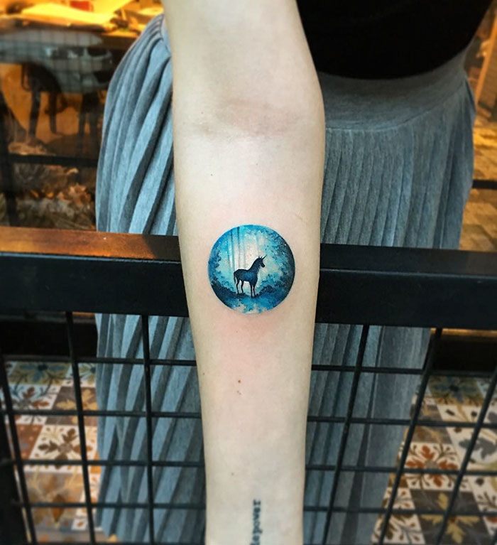 Miniature circle tattoo of a unicorn in a blue forest, created by Turkish artist Eva Krbdk.
