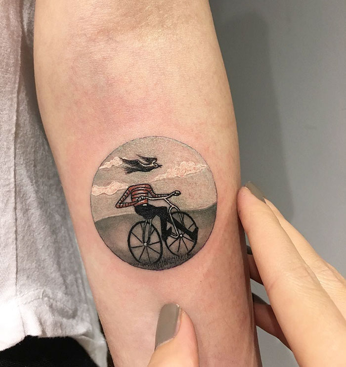 Miniature circle tattoo of a cyclist and bird by Turkish artist, on a person's arm, with finger pointing.