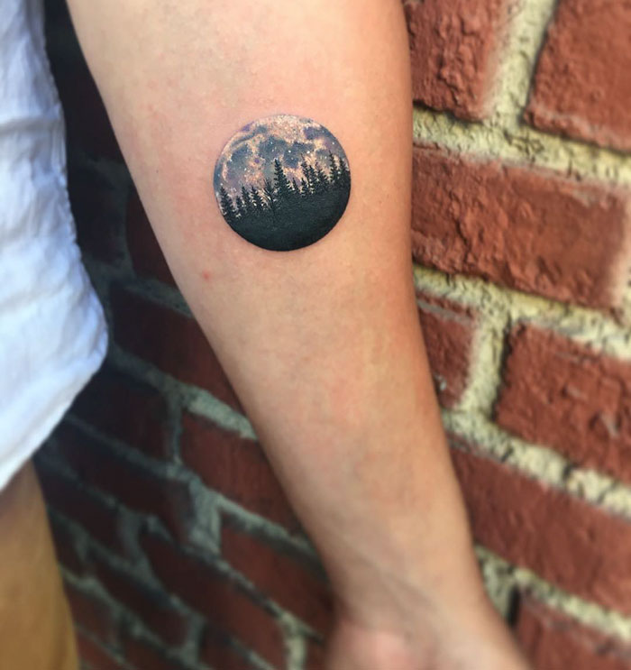 Miniature circle tattoo depicting a forest and night sky on a person's arm, created by artist Eva Krbdk.