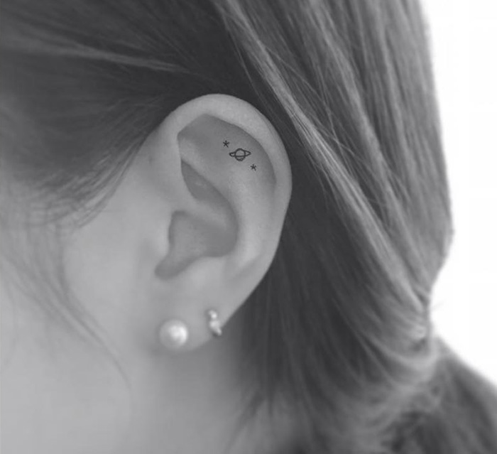 Minimalist tattoo by Korean artist on the upper ear, depicting a small planet with two stars nearby.
