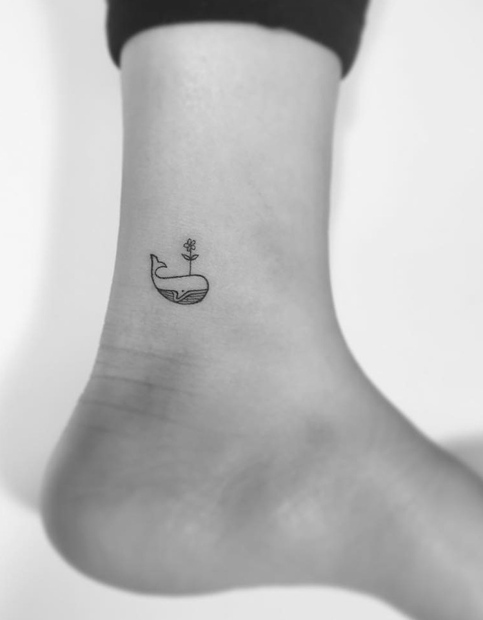 Minimalist whale tattoo with a small flower on an ankle by a Korean artist.
