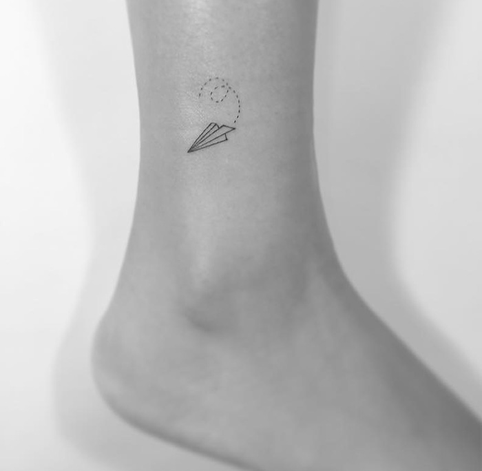 Minimalist tattoo of a paper airplane on an ankle, designed by a Korean artist.