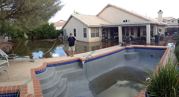 A man stands next to an empty swimming pool while the backyard is flooded, showcasing a job fail.