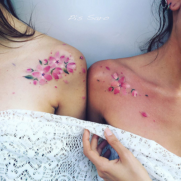 Two women with matching trio tattoos of pink flowers on their shoulders, showcasing delicate and artistic designs.