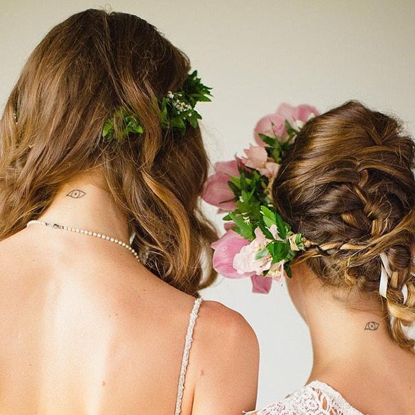 Two women with flower crowns and matching trio tattoos on their necks, symbolizing unity and friendship.
