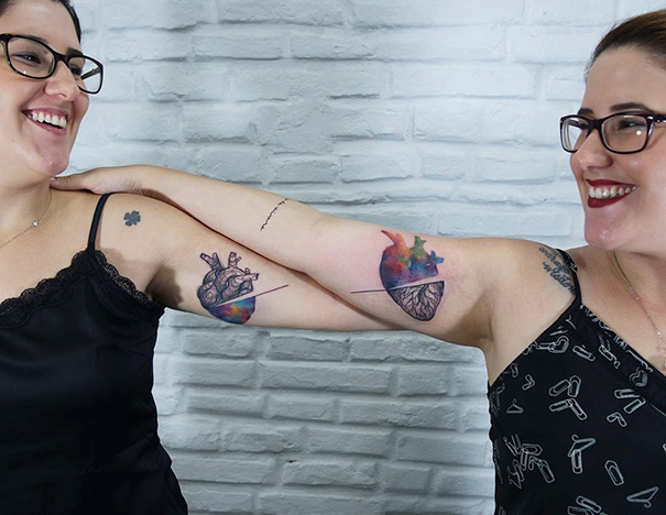 Two women showing trio tattoos on arms, featuring a heart and colorful designs, smiling at each other.