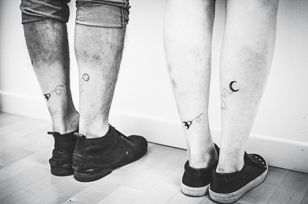 Black and white photo of two people showing trio tattoos on their calves, featuring a planet, key, and moon.