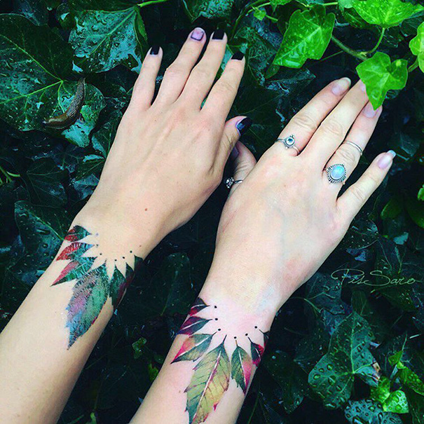 Trio tattoos of colorful leaves on two wrists with green foliage background.