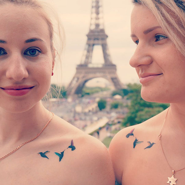 Two women with bird trio tattoos on shoulders, smiling near Eiffel Tower.