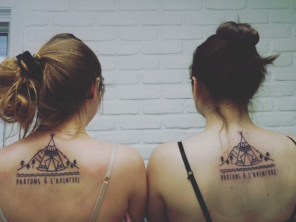 Two women with matching trio tattoos of a tent and phrase "Partons à l'aventure" on their upper backs.