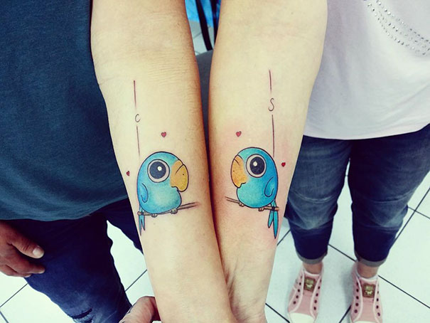 Two arms with matching parrot trio tattoos, each with a letter and red hearts, symbolizing unity and creativity.