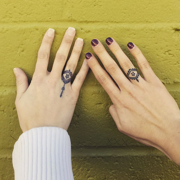 Two hands with geometric trio tattoos on ring fingers, against a yellow brick wall.