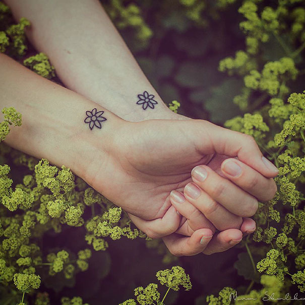 Two small flower trio tattoos on wrists amidst green foliage.
