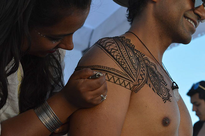 Man smiling while intricate henna tattoo is applied on his shoulder, showcasing the 'Menna' trend.