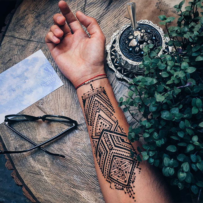 A man's arm with intricate henna tattoos resting on a table beside glasses and a potted plant.