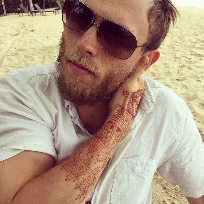 Man with intricate henna tattoos on forearm, wearing sunglasses and a white shirt at the beach.