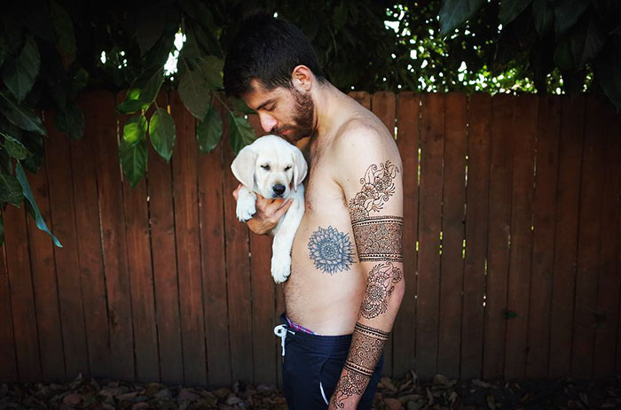 Man with intricate henna tattoos holds a puppy by a wooden fence.