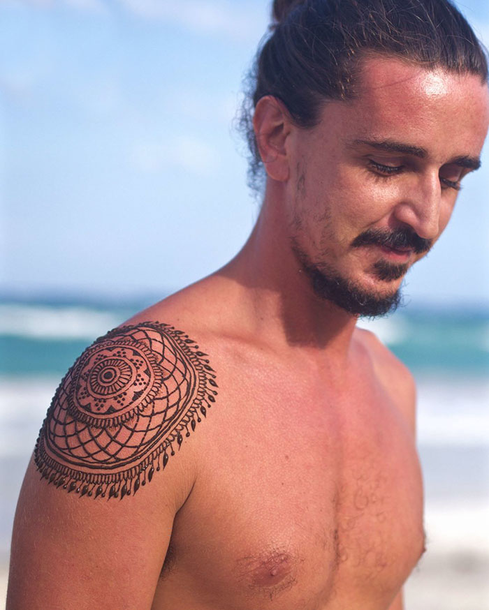 A man at the beach showcasing an intricate henna tattoo on his shoulder, part of the 'Menna' trend.