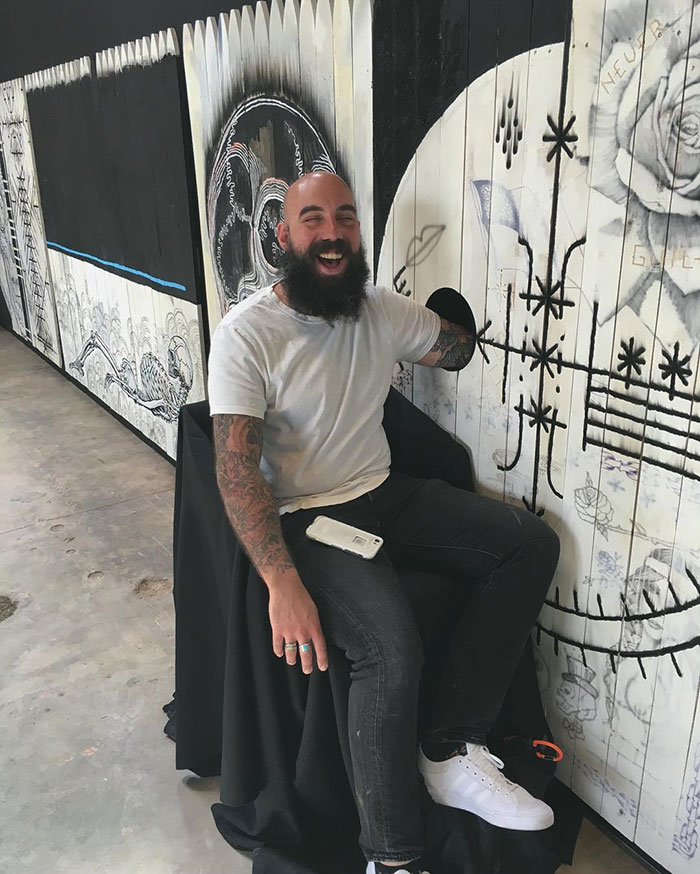 Person smiling while tattoo artist is tattooing through a hole in a decorated wall.