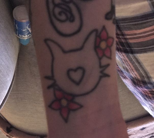 Cat tattoo design featuring a minimalist cat face with heart and flowers on an arm.