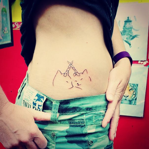 Minimalist cat tattoo on a person's side, featuring two cat faces with a braid, in a vibrant tattoo studio.