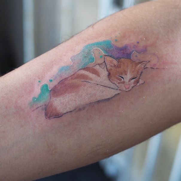 Watercolor-style cat tattoo on an arm, featuring a sleeping cat with colorful background. Perfect for cat person expression.