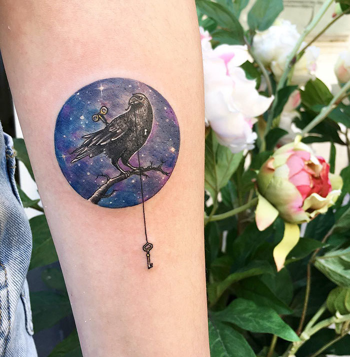 Small bird tattoo featuring a cosmic background and a key, inked on forearm near flowers.