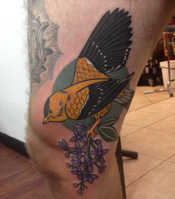 Colorful small bird tattoo holding purple flowers on a person's leg.