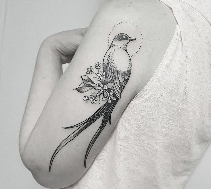 Small bird tattoo with floral accents on upper arm, highlighting intricate design and elegance.