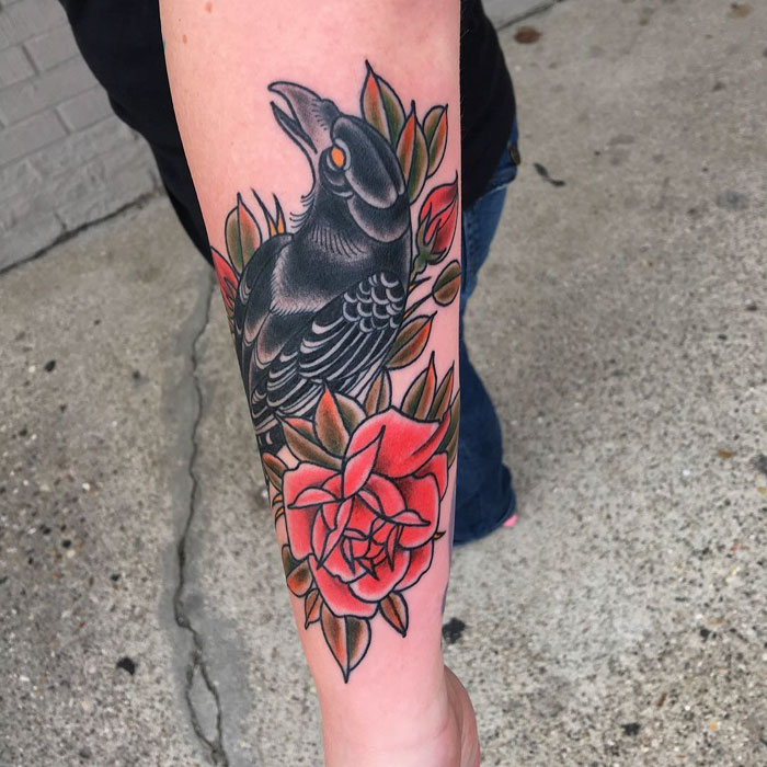 Unique small bird tattoo featuring a black bird with vibrant red roses on the forearm.