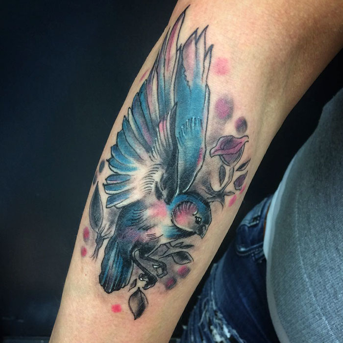 Colorful small bird tattoo on forearm with floral accents.