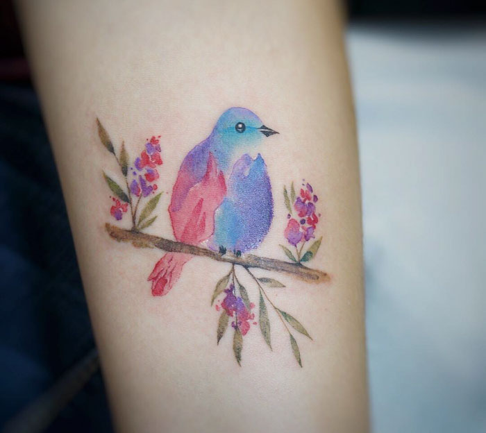 Colorful small bird tattoo on a branch with flowers, showcasing intricate ink artistry.