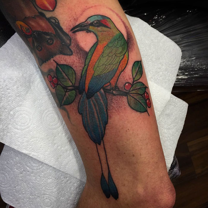 Colorful small bird tattoo with green wings and orange accents on a branch, inked on an arm.