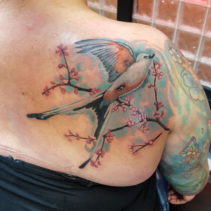 Small bird tattoo with pink blossoms on shoulder, featuring vibrant colors and intricate design.
