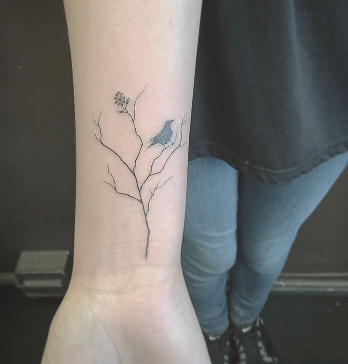 Small bird tattoo on forearm featuring a bird perched on a branch with a flower detail.