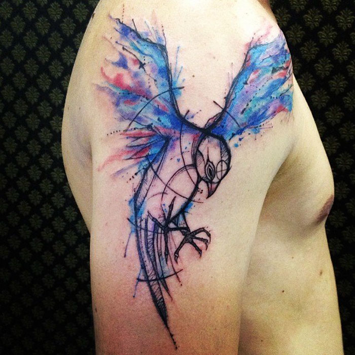Abstract small bird tattoo with vibrant watercolor on upper arm.