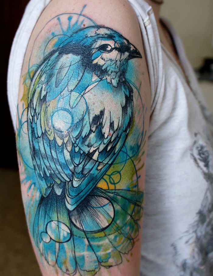 Colorful small bird tattoo on arm with abstract watercolor design.